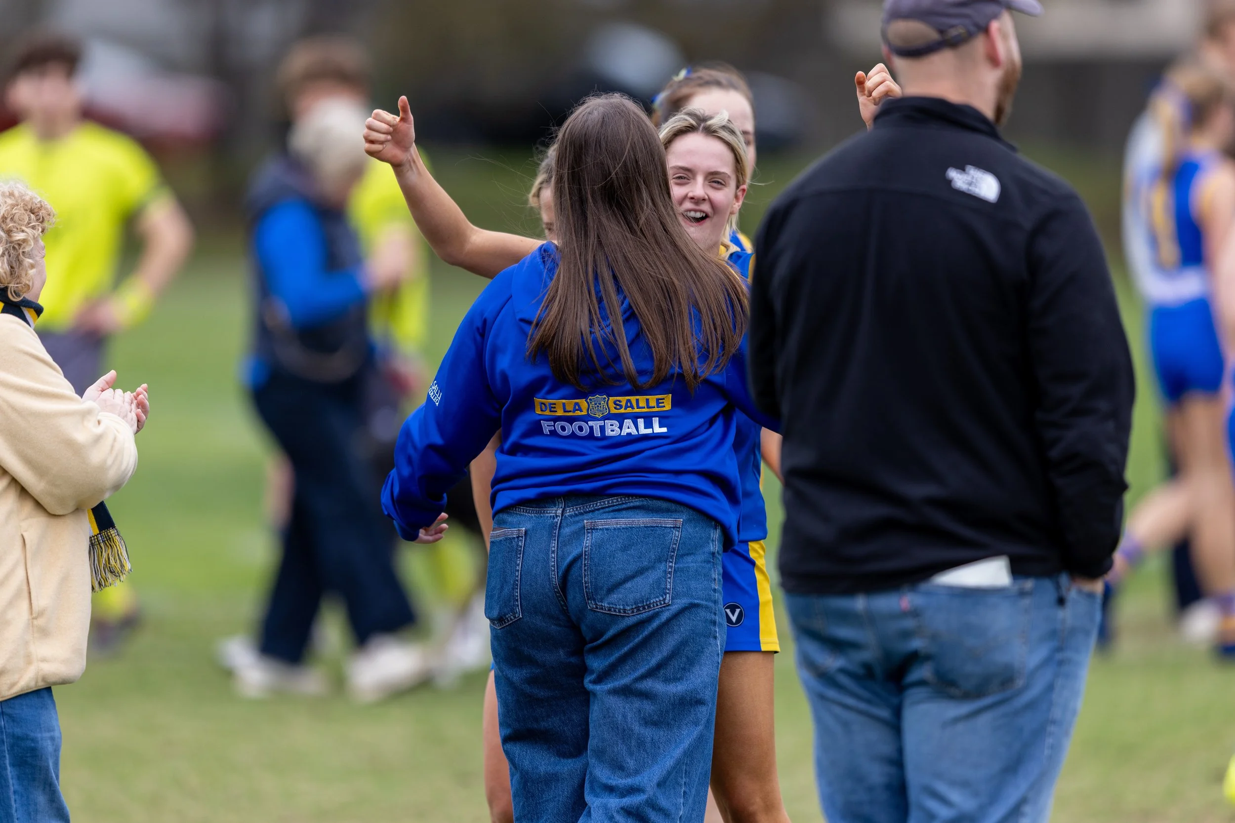 Group of children and adults outdoors on a grassy field, engaged in a sporting event. The children are wearing blue and yellow sports uniforms, with one girl smiling and holding her arm up in a celebratory gesture. Other children and an adult are seen clapping and talking, with a background of blurred spectators and trees.