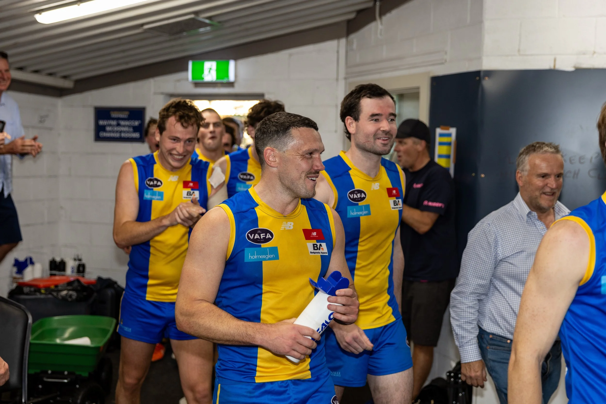 Group of male De La Salle Footballers in yellow and blue uniforms celebrating in a locker room, with some smiling and holding a white water bottle.