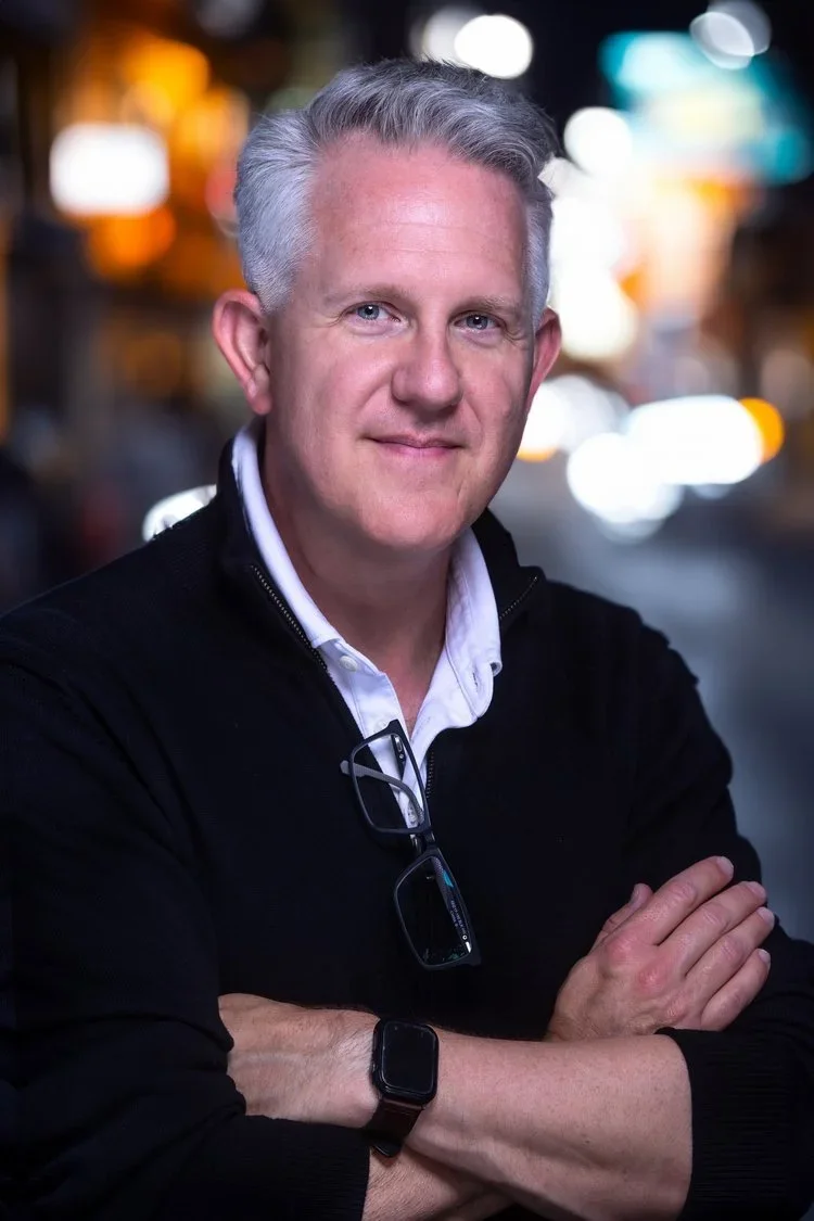 A middle-aged man with gray hair and blue eyes stands outdoors at night, wearing a black jacket over a white collared shirt. He has sunglasses hanging from his shirt, a smartwatch on his left wrist, and a pair of glasses clipped to his shirt. His arms are crossed, and he is smiling slightly, with a city street and blurred bright lights in the background.