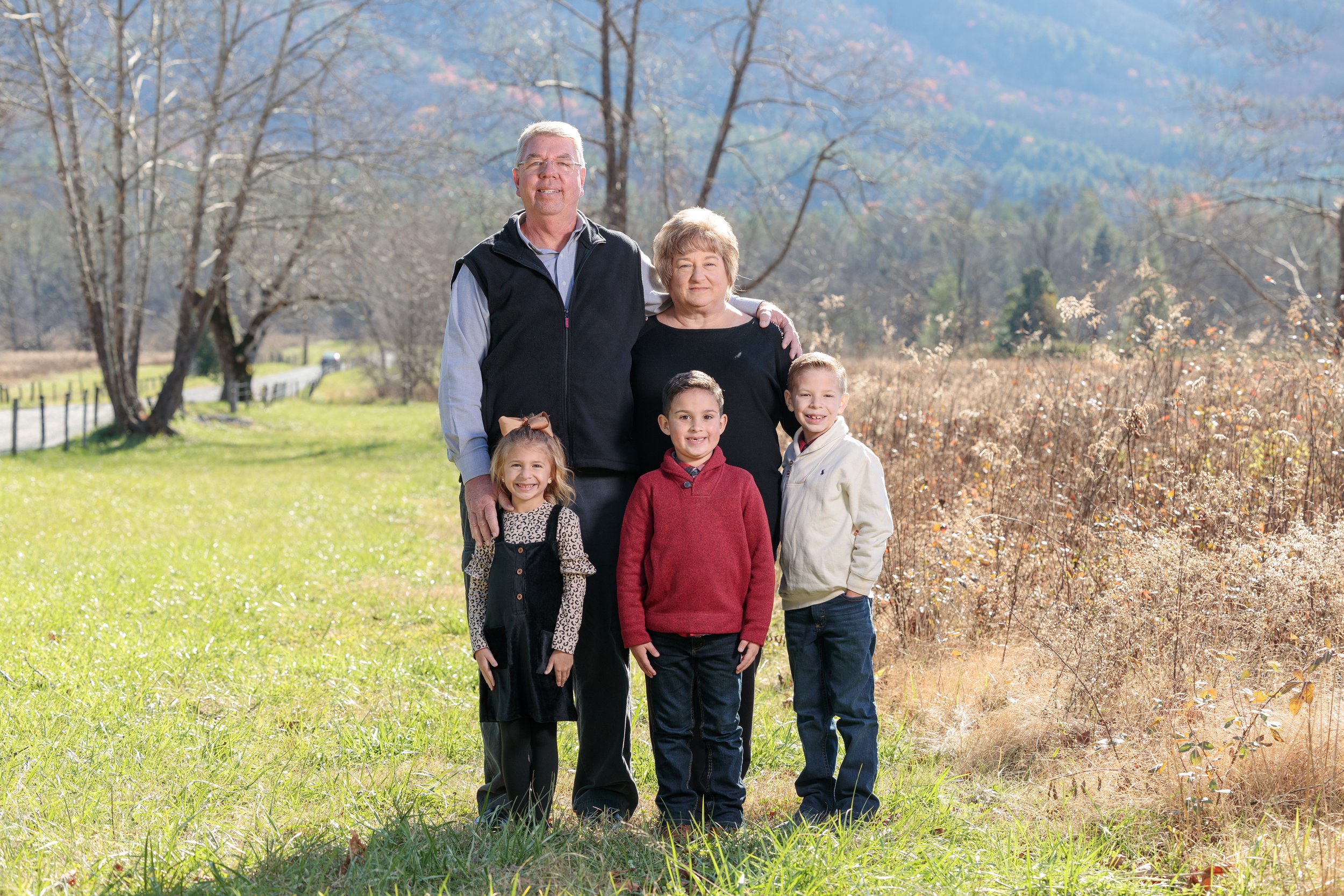 Grandparents posing with their three young grandchildren in a sunny Cades Cove meadow with mountain views in the background.