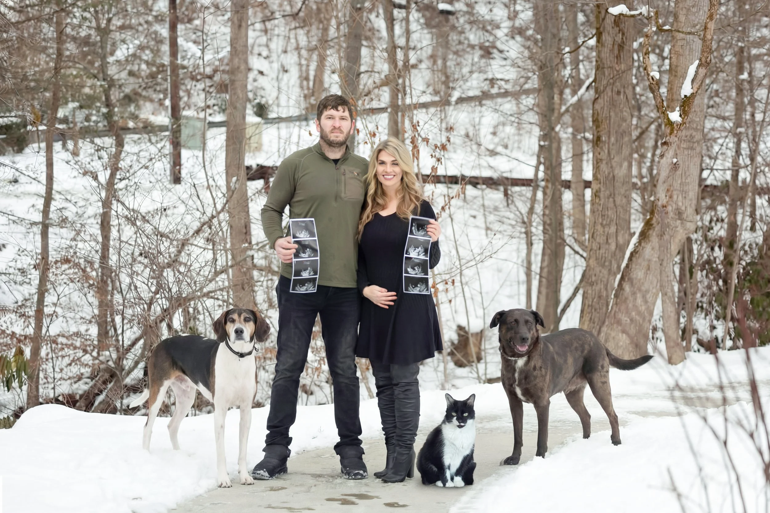 A couple with three dogs and a cat standing outdoors on snow-covered ground in a forest during winter. The woman is pregnant and holding ultrasound pictures, while the man is next to her, both smiling.