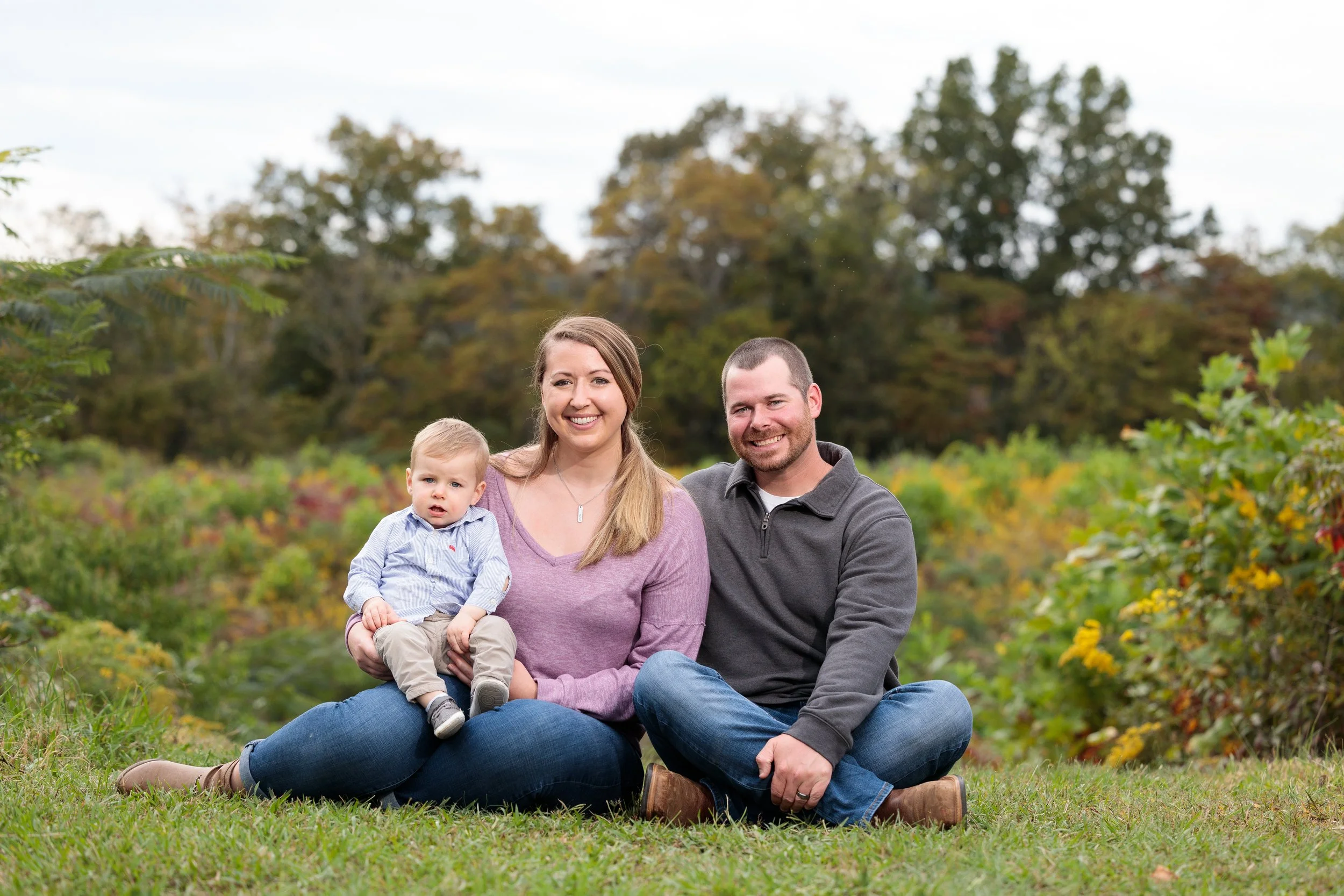 A happy family of three with a woman, a man, and a young boy sitting on grass in a park with trees in the background.