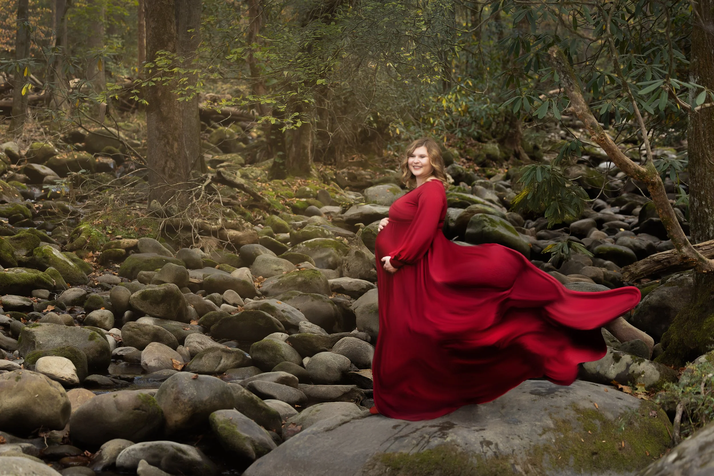 A woman wearing a long, flowing red dress sitting on rocks in a wooded area with forest trees and moss-covered stones, smiling.