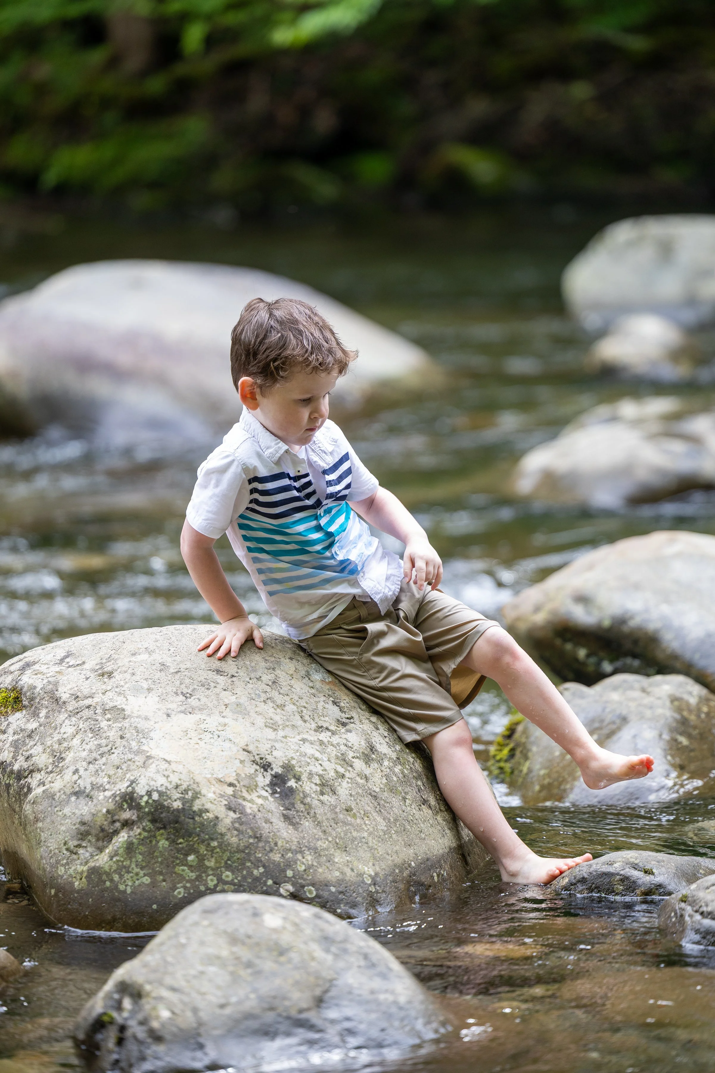 Young boy sitting barefoot on a large river rock with his feet dangling in the mountain stream.
