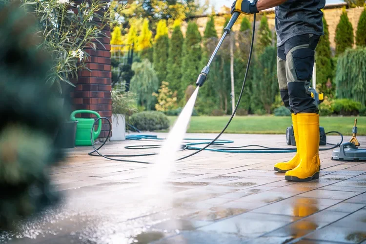 Person wearing yellow rubber boots power washing a tiled outdoor patio in a garden.