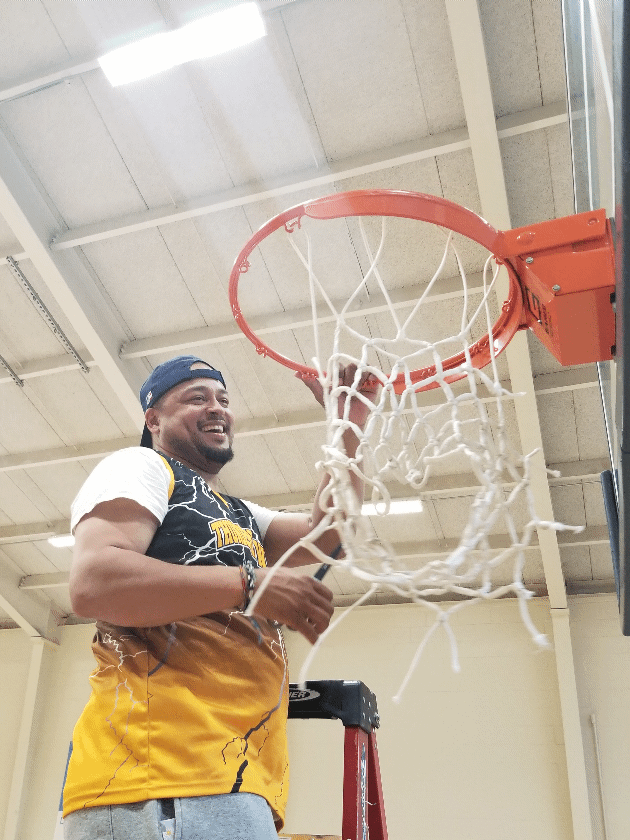 A man wearing a basketball jersey and a backward cap is smiling while hanging on a basketball hoop in a gymnasium.