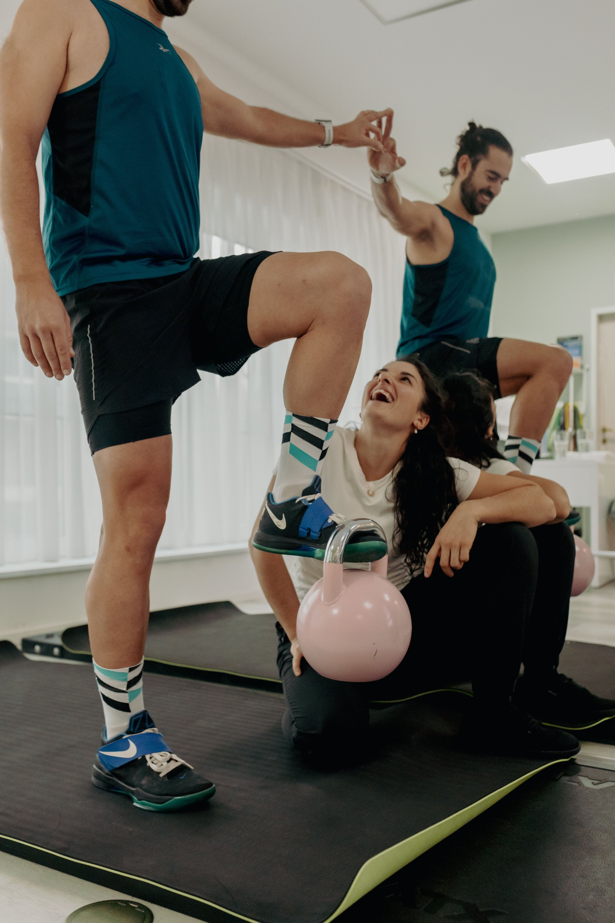 Zwei Männer trainieren in einem Fitnessstudio, eine Person macht eine Kniebeuge, die andere unterstützt und zeigt die Hand des Partners. Eine Frau liegt auf dem Boden und hebt den Fuß in einer Kniebeuge, während sie lachend das Gesicht zur Kamera dreht. Es gibt ein rosa Kettlebell und ein Yogamatte im Hintergrund.