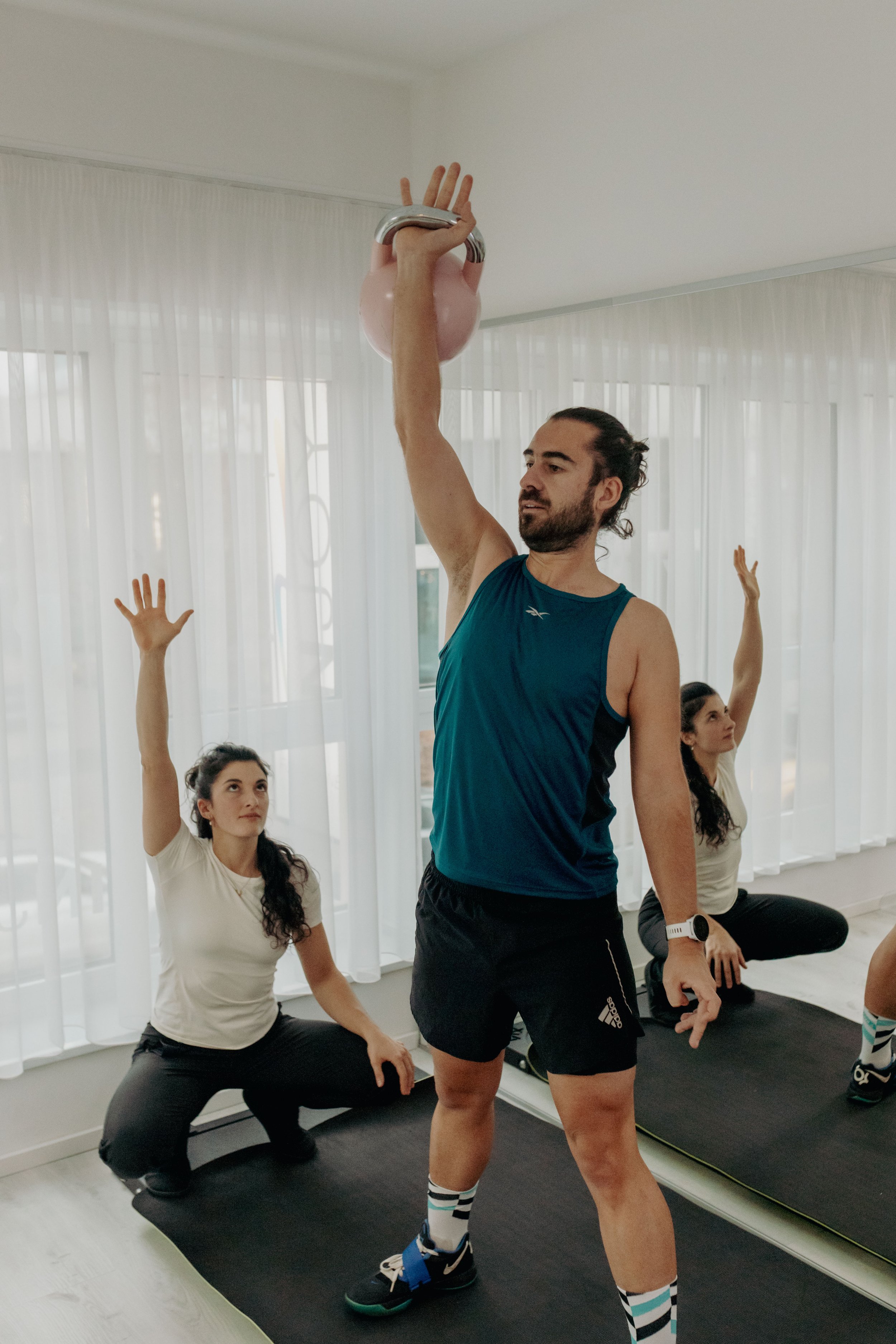 Ein Mann trainiert mit einer Kettlebell, während drei Frauen in der Nähe auf einer Yogamatte sitzen und die Arme heben, in einem hellen Raum mit großen Fenstern und weißen Vorhängen.