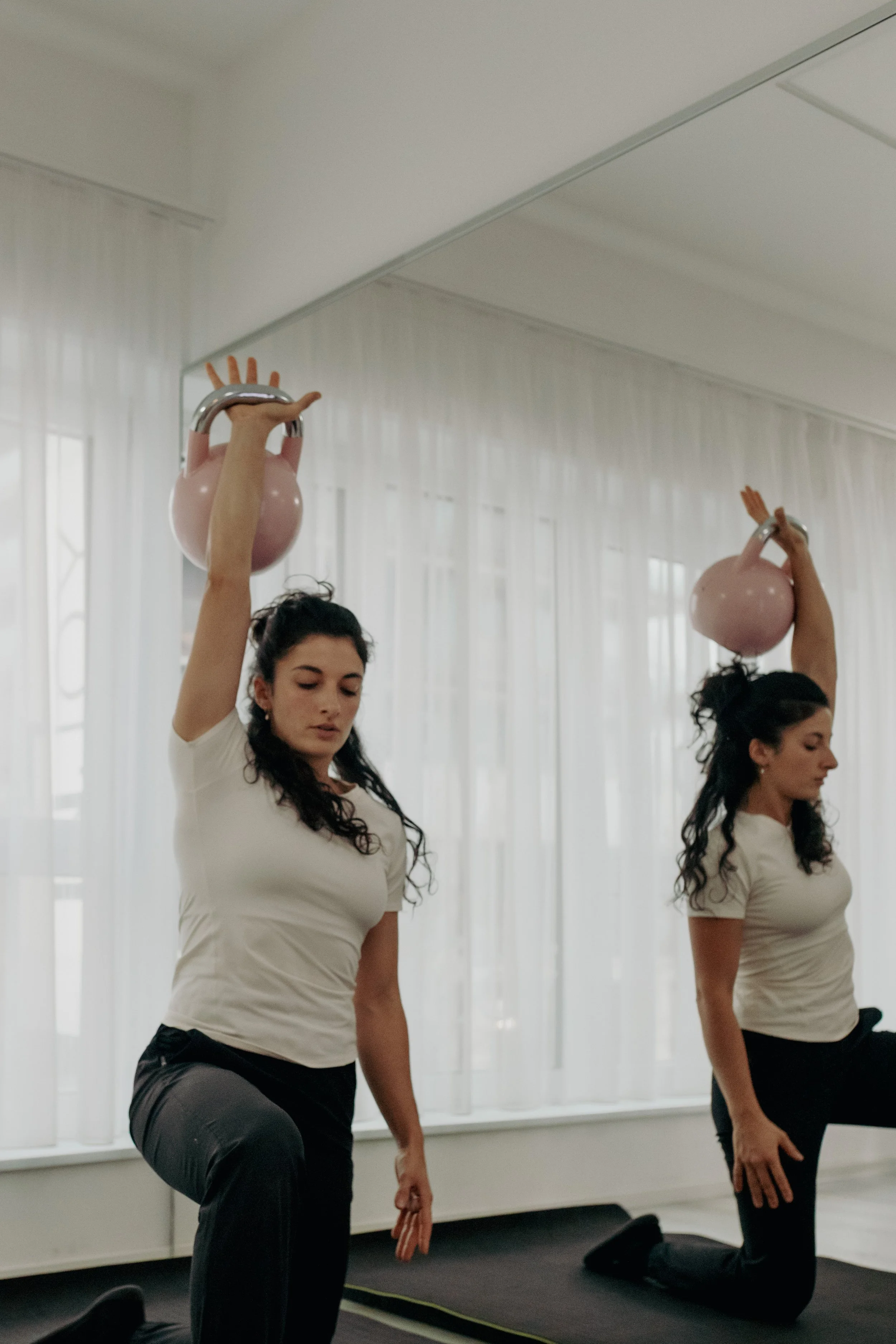 Drei Frauen beim Yoga-Übung in einem Raum mit großen Fenstern und weißen Vorhängen. Die Frauen tragen weiße T-Shirts und schwarze Hosen, eine hält ein pinkes Gewicht über dem Kopf, die andere befindet sich in einer geknietten Position.