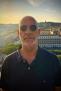 Ein Mann trägt eine dunkle Sonnenbrille und ein dunkelblaues Polo-Shirt, im Hintergrund eine Stadt bei Sonnenuntergang mit Gebäuden und Bäumen.