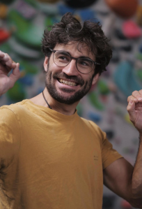 Ein lachender Mann mit Brille, grauem lockigem Haar und Bart, trägt ein gelbes T-Shirt und scheint im Freien unterwegs zu sein.