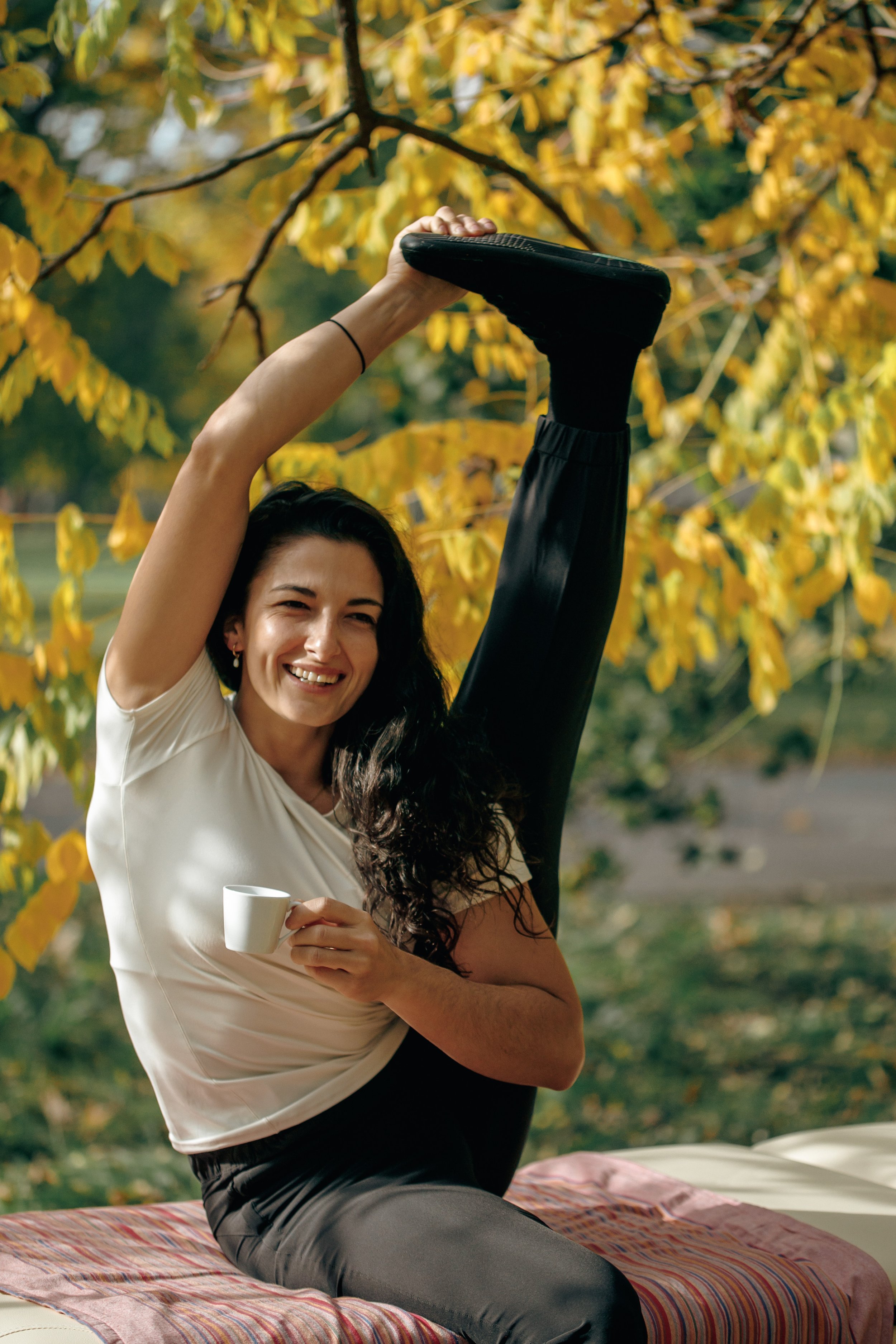 Eine lachende Frau hält ein Hüftbein nach oben, um die Flexibilität zu zeigen, während sie im Freien bei gelben Herbstblättern sitzt. Sie trägt eine weiße Bluse, schwarze Leggings und einen schwarzen Haargummi. Sie hält eine Tasse in ihrer linken Hand, während sie auf einer bunten Decke sitzt.