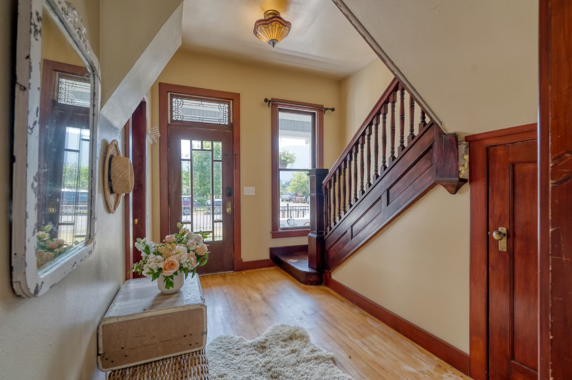 Entryway with wooden door and window, staircase with wooden railing, floral arrangement on table, mirror on wall, and cozy rug on hardwood floor.