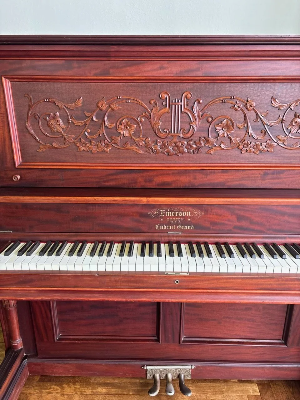 A vintage upright piano with ornate wooden carvings and a logo that reads 'Emerson.' The piano has a closed top and a keyboard with black and white keys. The wood has a dark, polished finish.