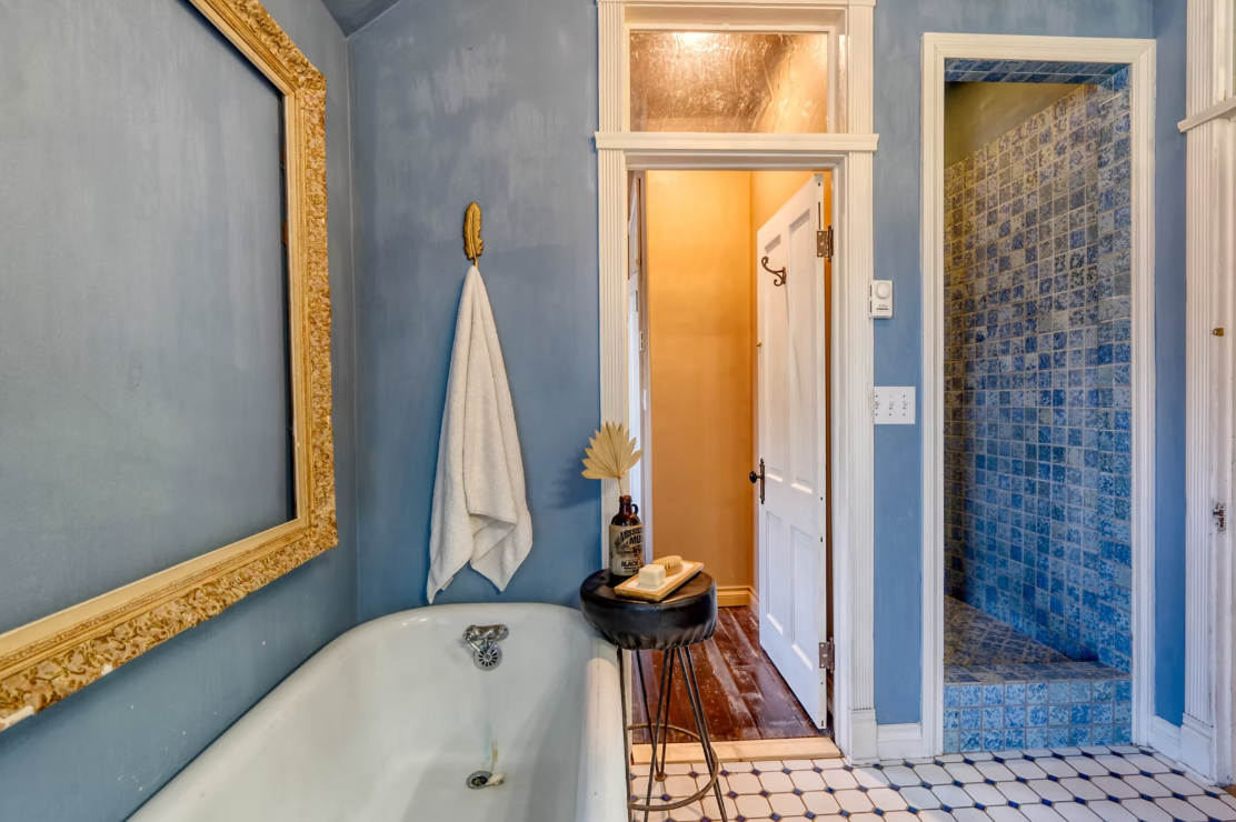 A vintage bathroom featuring a clawfoot bathtub, a small side table with soap and a decorative bottle, a wall hook with a white towel, a large ornate mirror, and a walk-in shower with blue mosaic tiles. The walls are painted blue, and the floor has black and white tile patterns.
