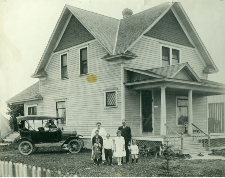 A vintage black and white photo of a large two-story house with a front porch, and seven children and one adult standing in front. A car is parked near the house, and a dog is also visible near the porch.