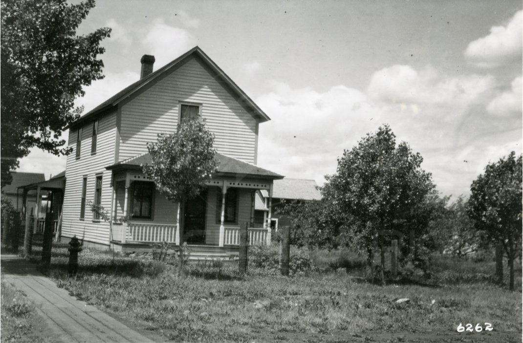 Black and white photo of a two-story American house with a front porch, surrounded by trees, and a fence in the front yard.