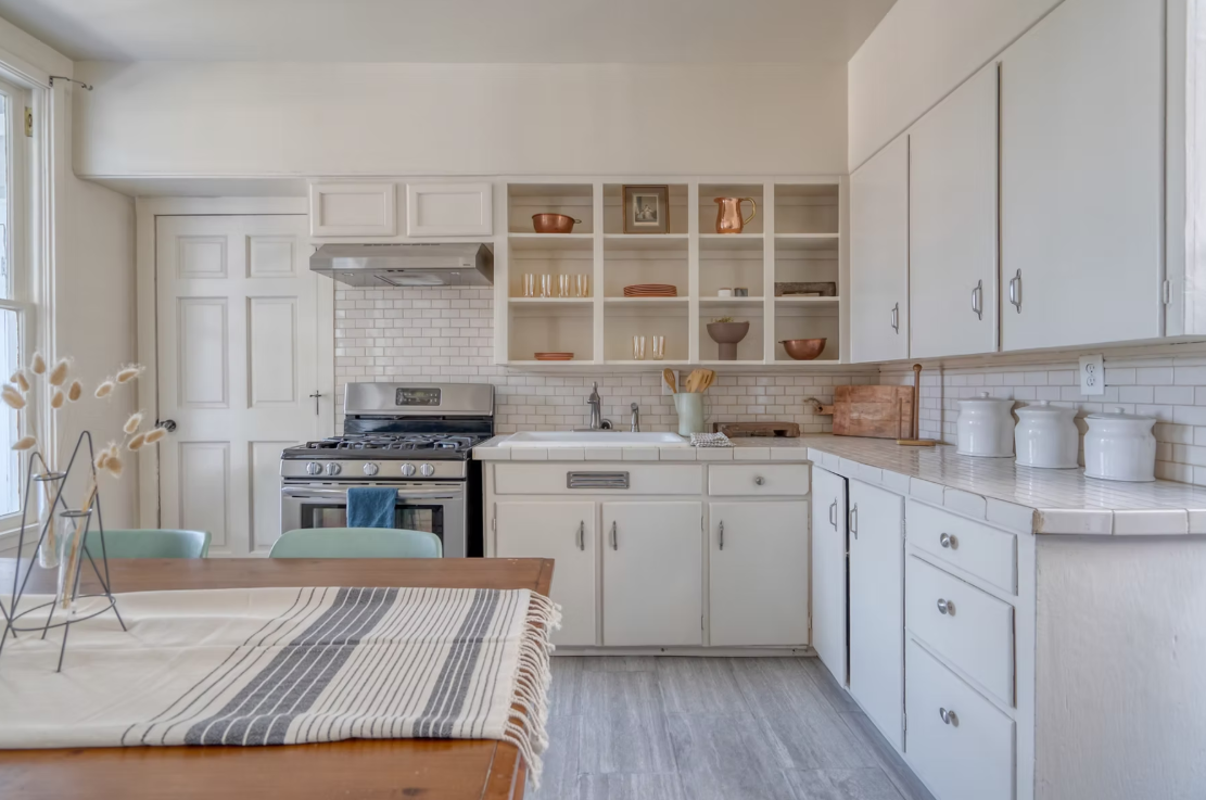A cozy kitchen with white cabinets, open shelving displaying dishes and decorative items, a stainless steel oven, a white brick backsplash, a farmhouse sink, and a wooden dining table with a striped table runner.
