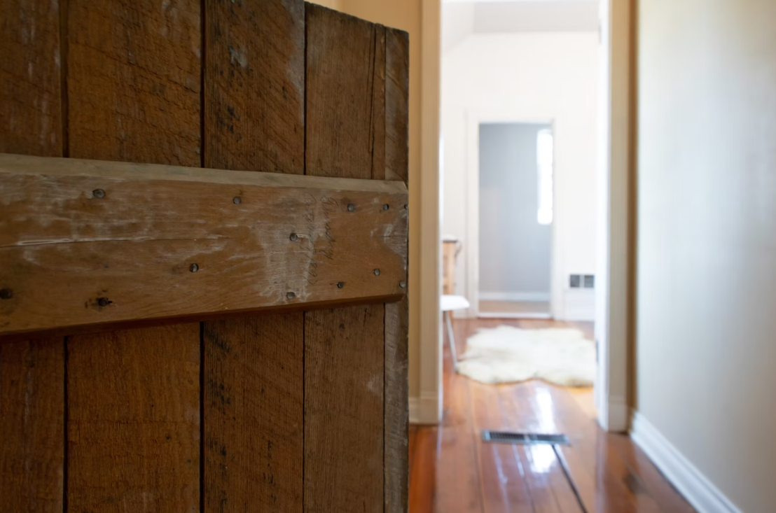 Partial view of a rustic wooden door and a hallway with hardwood flooring, a cozy rug, a chair, and a window at the end of the hall.