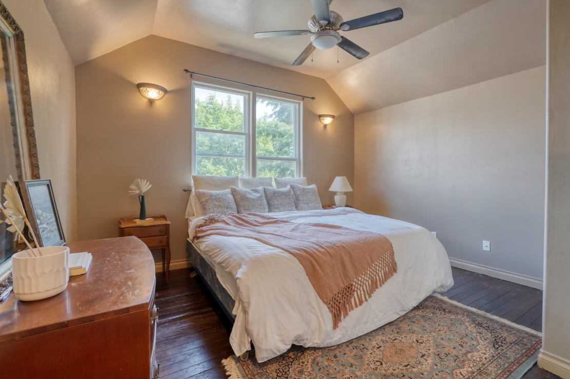 A cozy bedroom with a large bed covered in white and beige bedding, positioned against a wall with two windows showing green trees outside. The room has a ceiling fan, wall sconces, a wooden dresser with decorative items, a bedside table with a lamp, and a patterned area rug on dark wood floors.