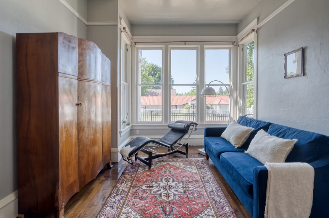 Living room with large bay window, wooden wardrobe, black chaise lounge, blue sofa with white pillows, patterned area rug, wall-mounted reading lamp, and framed artwork