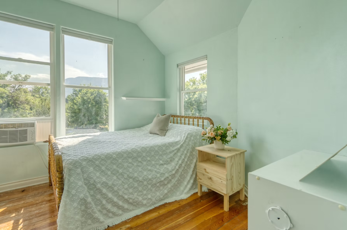 A minimalistic bedroom with mint green walls, a wooden bed with a white textured bedspread and a beige pillow, a small wooden nightstand with a flower vase, and large windows letting in natural light revealing a view of trees and mountains.