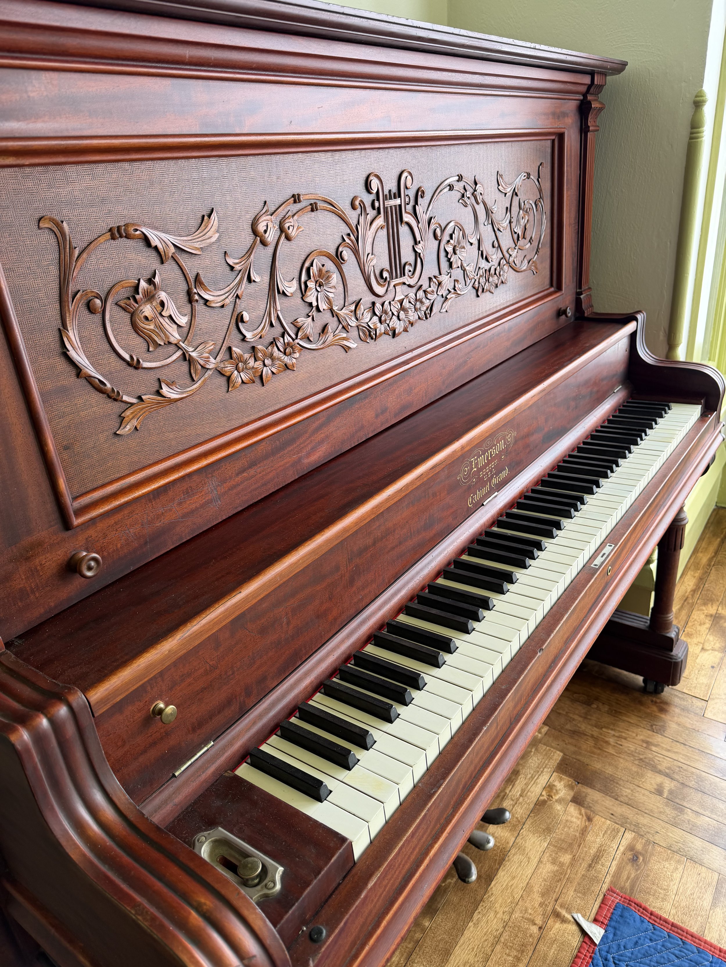 A vintage upright piano with ornate wood carvings on the front panel, black and white keys, and brass pedals, situated on a wooden floor next to a yellow wall.