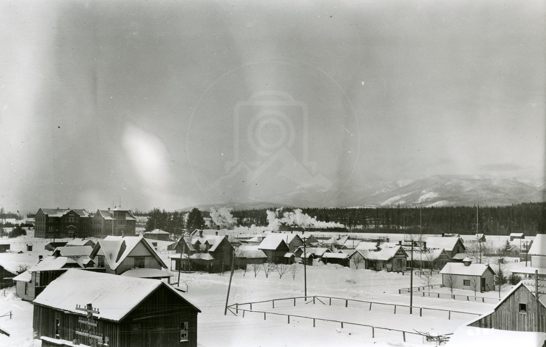 A black and white photograph of a snowy town with houses and a large building in the background, with snow-covered mountains in the distance.