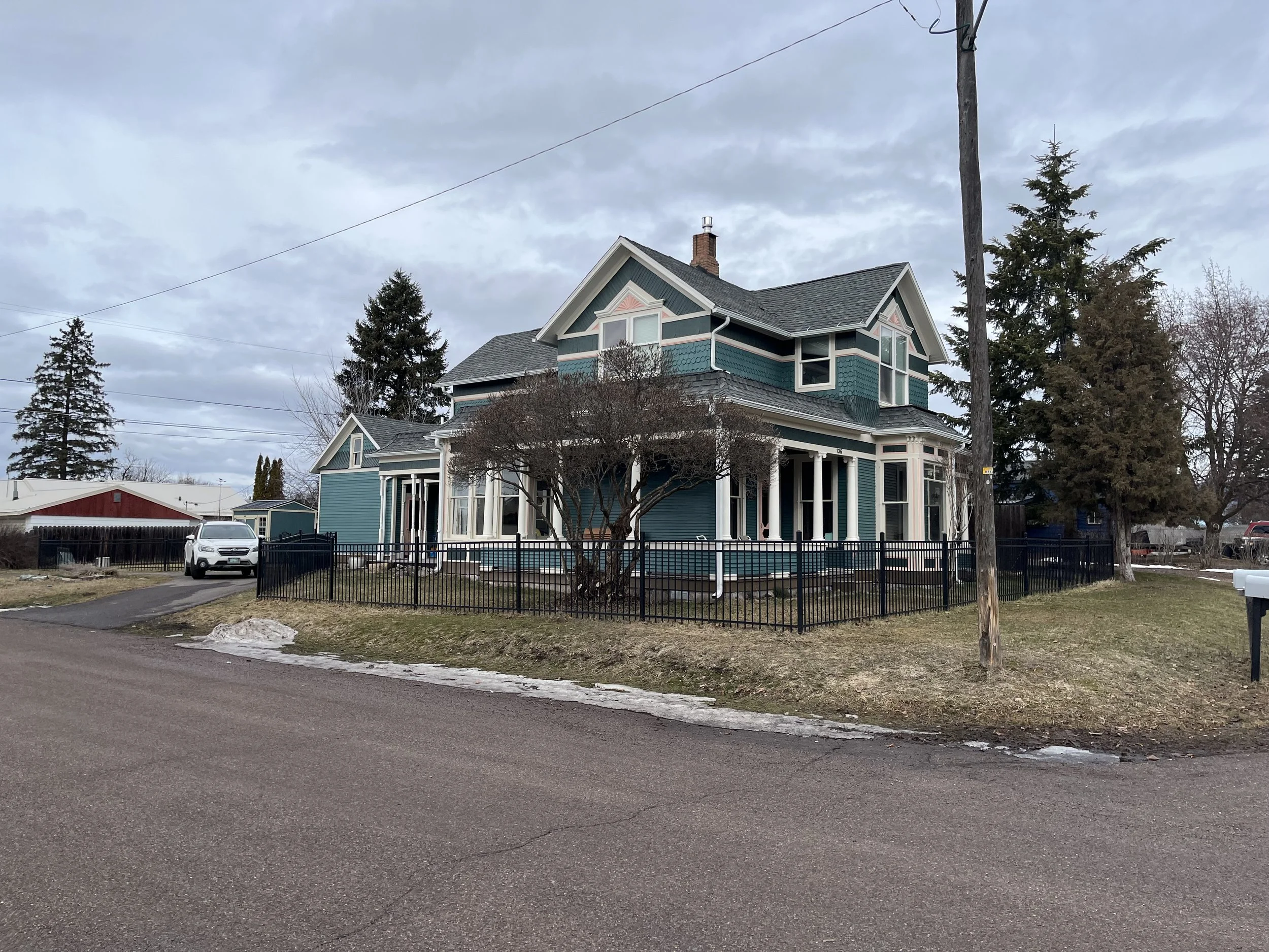 A two-story blue house with white trim and decorative shingles, surrounded by trees and a metal fence, with a parked white vehicle nearby and a cloudy sky overhead.