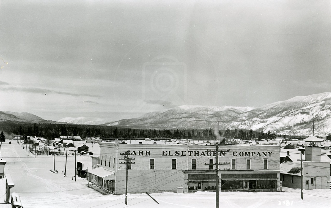 Black and white photo of a snowy town with mountains in the background, featuring buildings and utility poles.