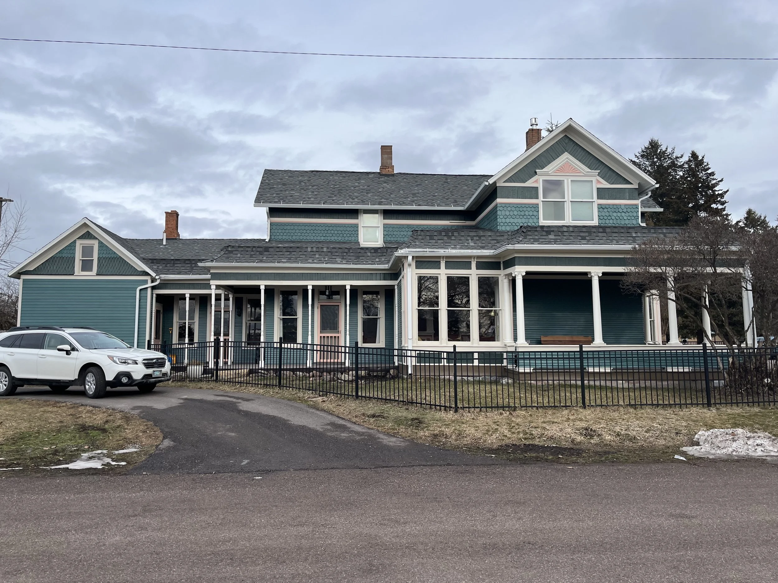 A large, two-story house with teal siding, white trim, and a gray shingled roof. The house has multiple windows, including a prominent bay window, and several chimneys. A black metal fence surrounds the front yard, which has a pathway leading to the front porch. There is a white car parked in the driveway, and some patches of snow on the ground. The sky is overcast with gray clouds.