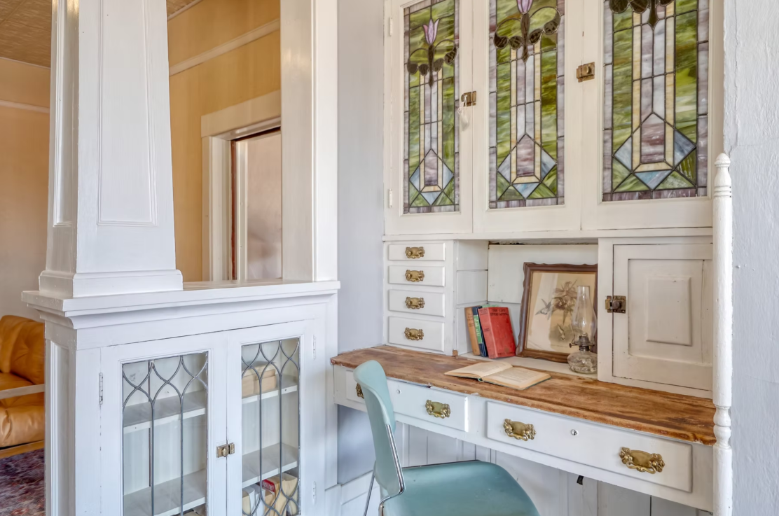 White built-in cabinet with stained glass doors and a wooden desk surface, with books, a framed picture, and an oil lamp on it, in a room with white walls.