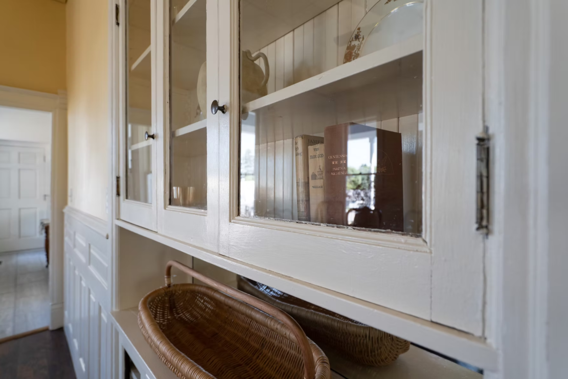 A white wooden cabinet with glass doors, containing plates and books inside, and a wicker basket below.