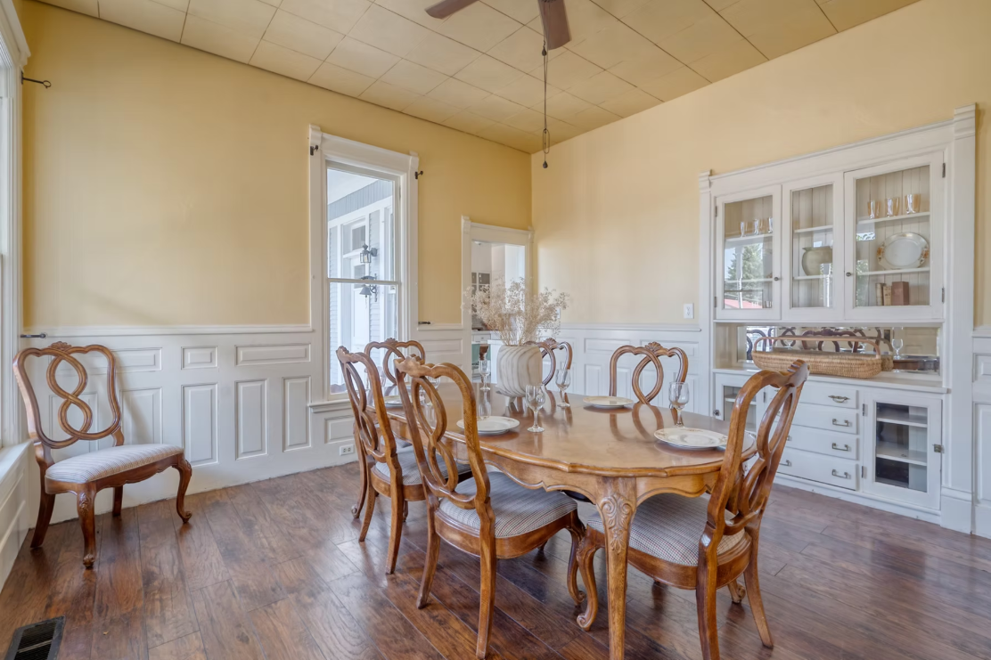 A cozy dining room with a round wooden table set with plates and glasses, surrounded by wooden chairs, with a china cabinet displaying dishes and glasses, and a large window letting in natural light.