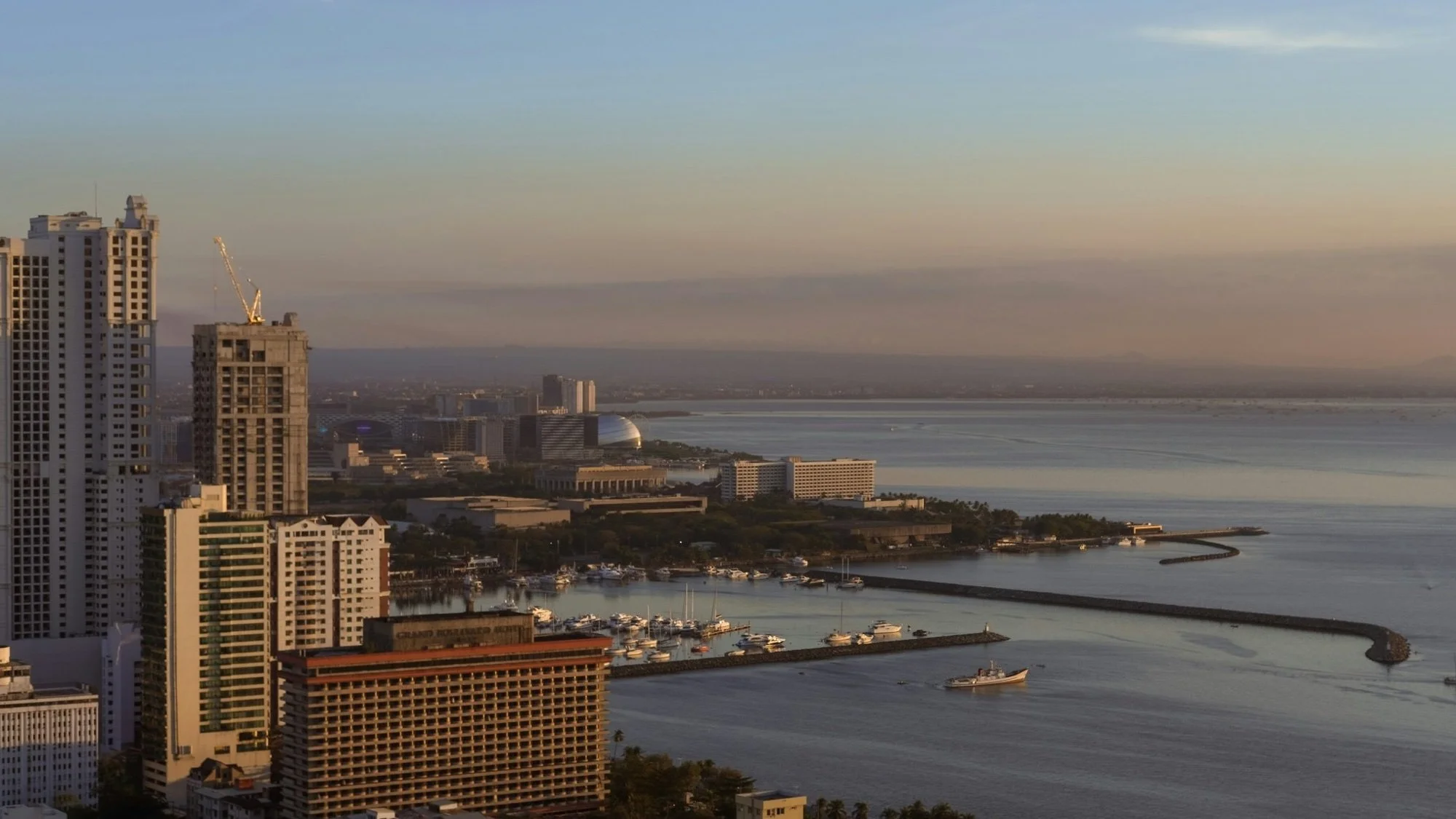 Manila Bay at golden hour, with a marina, a curving breakwater and calm water beneath a soft sky.