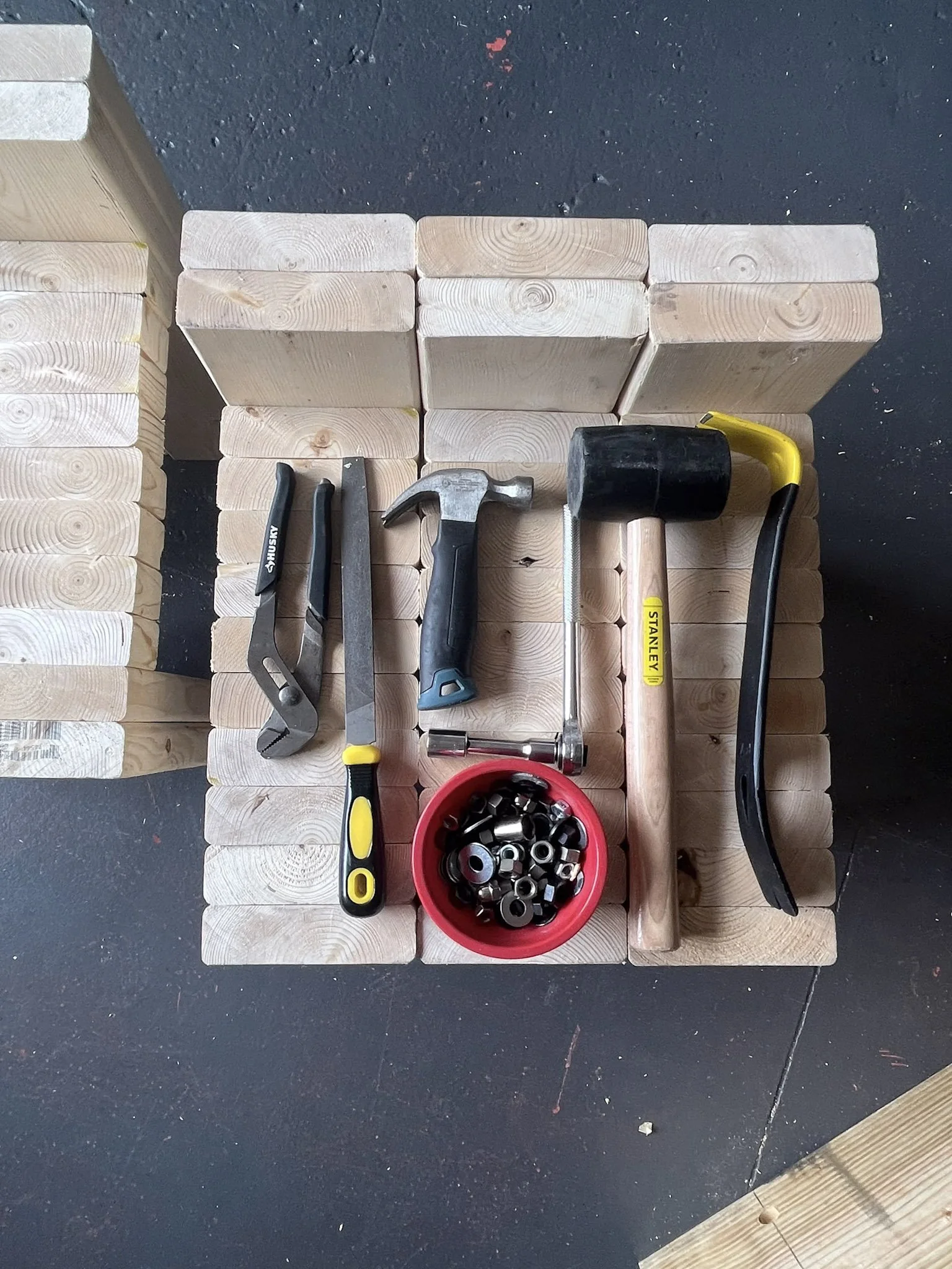 Wooden blocks arranged on a black surface with various tools, a hammer, a screwdriver, a wrench, and a container of nuts and bolts.