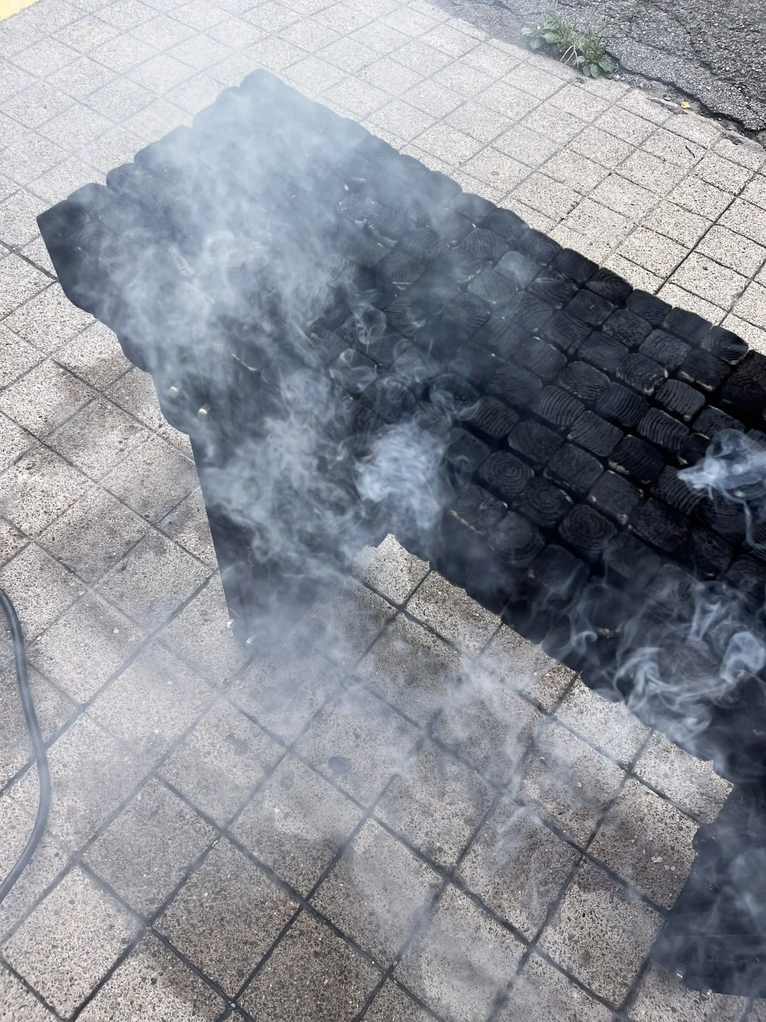 Close-up of a charcoal barbecue grill with smoking hot coals on a paved sidewalk, with some small plants along the edge.
