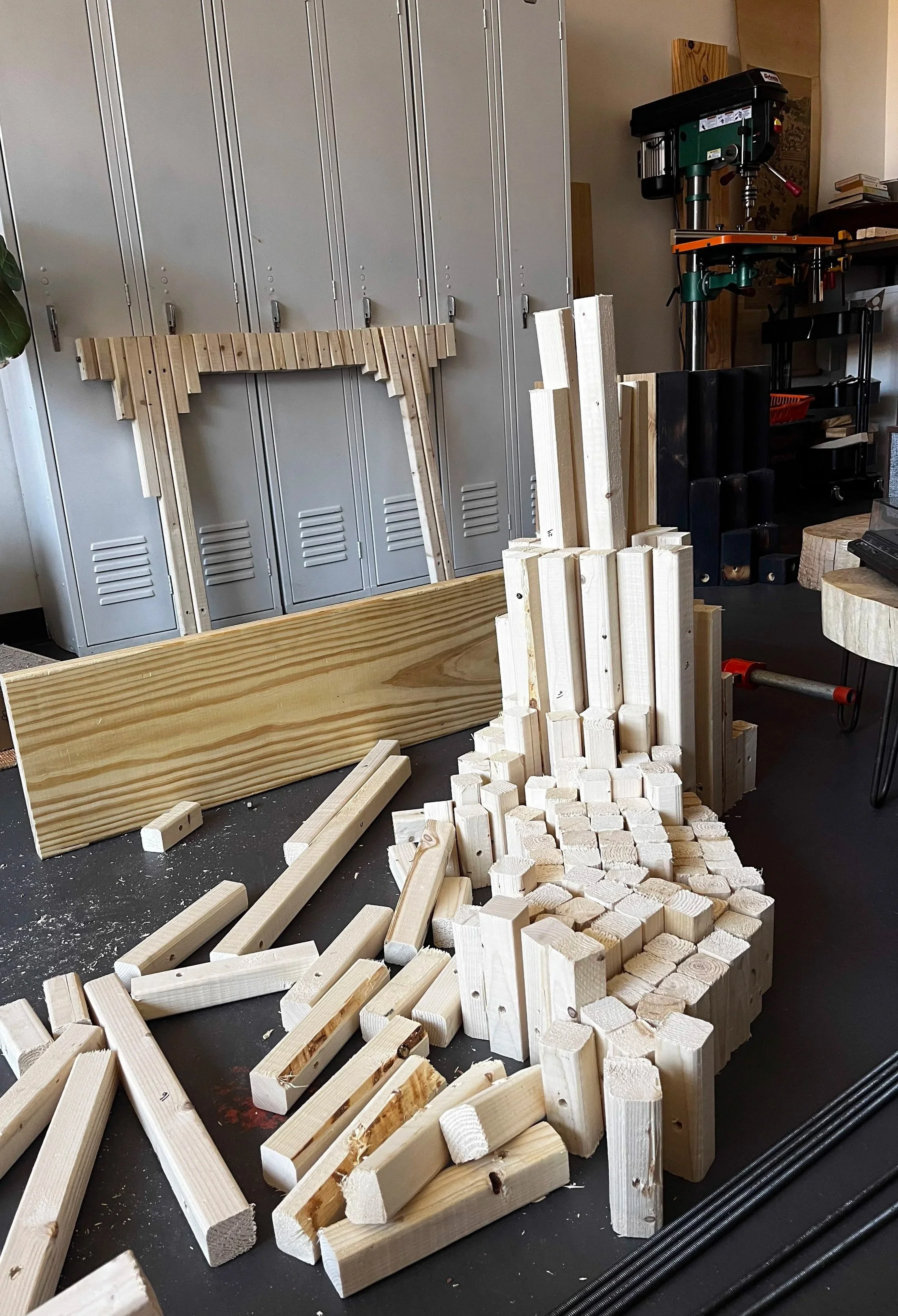 Woodworking workshop with various unfinished wooden pieces, a drill press, and storage lockers in the background.