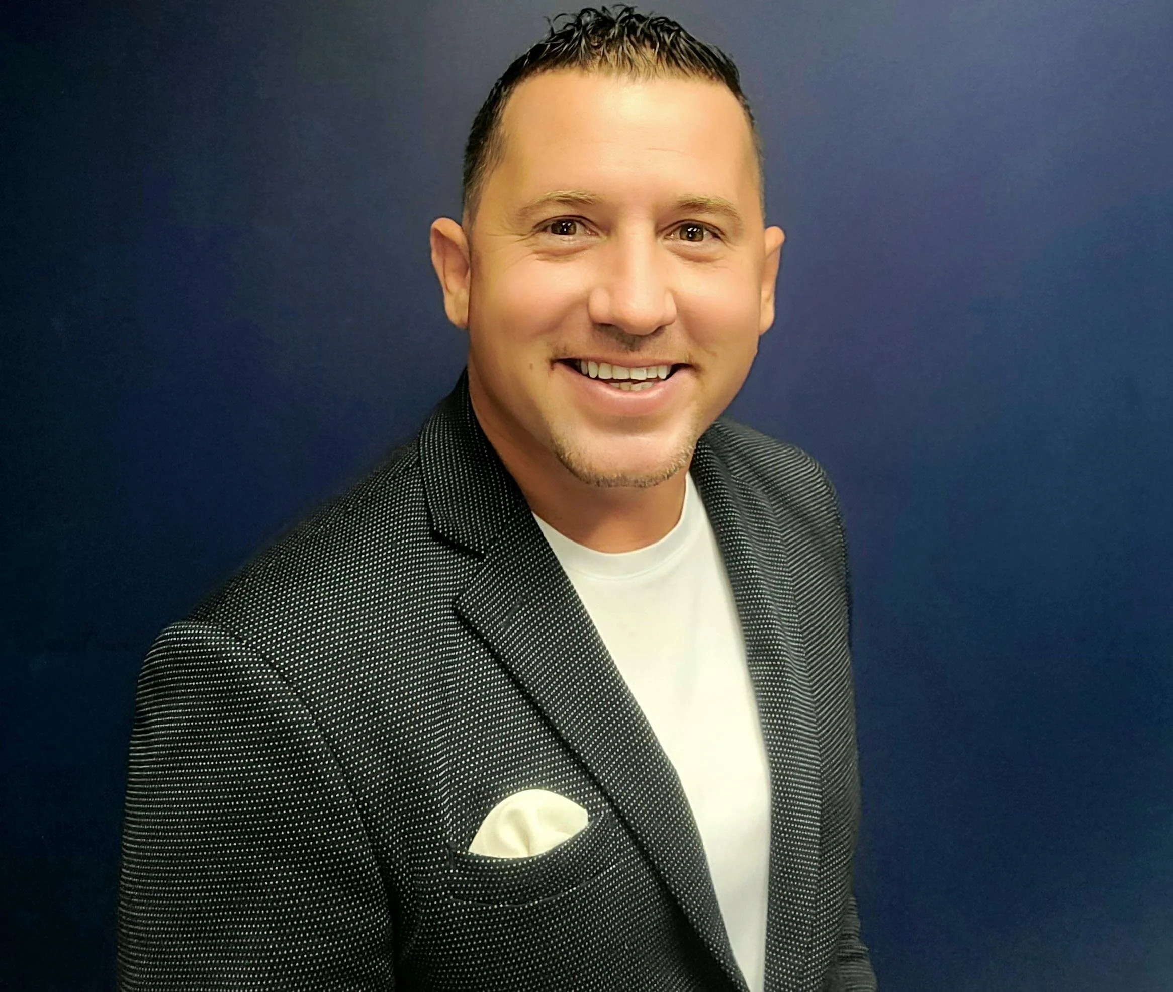Close-up of a smiling man in a black dotted blazer and white shirt, standing against a dark blue background.