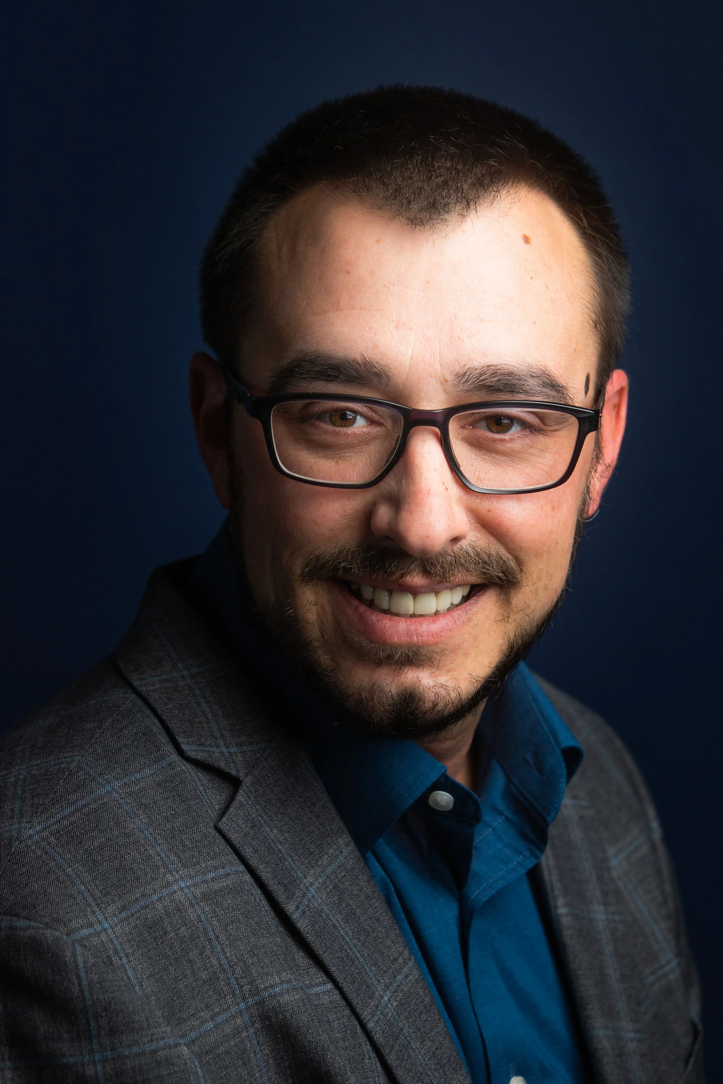A smiling man wearing glasses, a dark blue shirt, and a gray blazer with a checkered pattern.