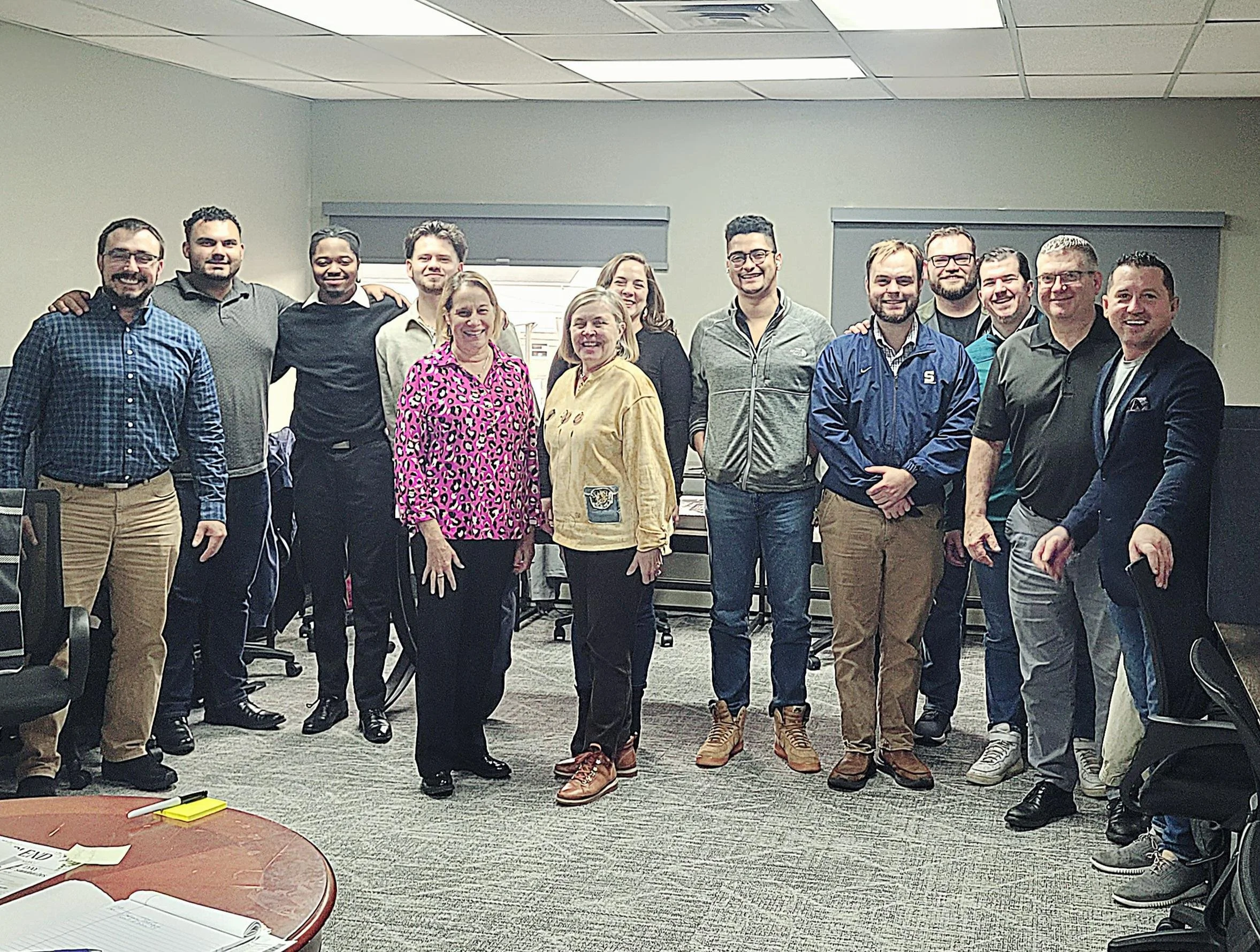 A group of fifteen people standing together in an office conference room, smiling at the camera.