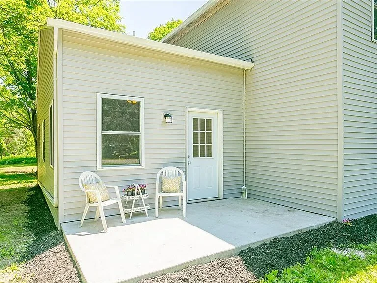 Small outdoor patio area with two white plastic chairs, a small table with flower pot, near a beige house with a door and window, green grass and trees in the background.