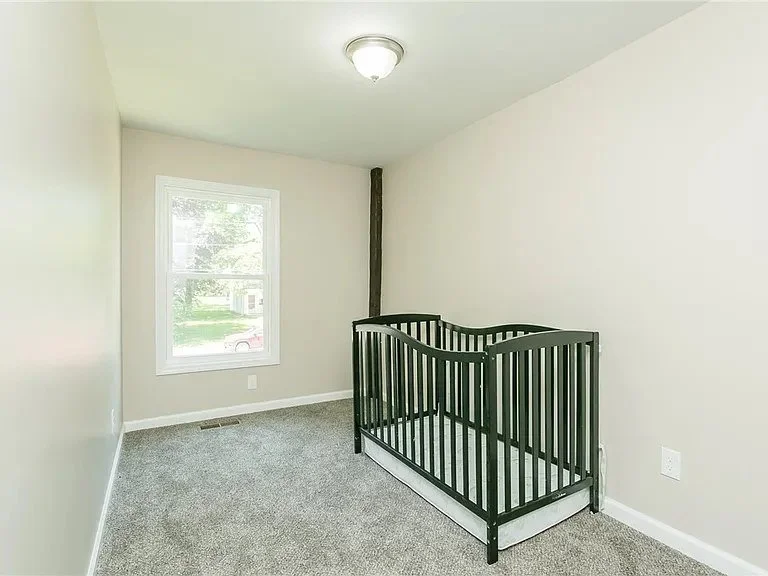 Empty nursery room with beige walls, a window, gray carpet, and a black crib.