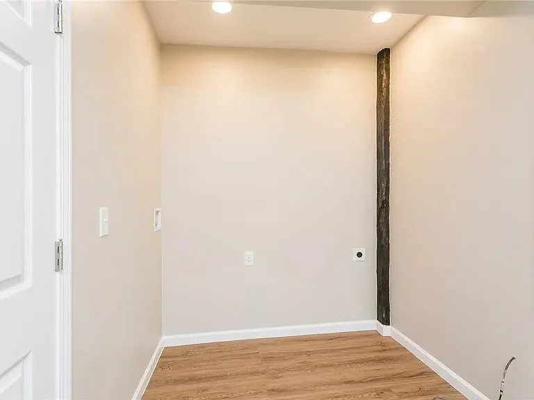 Empty room with beige walls, wood flooring, and a dark vertical beam on the right wall. There are recessed ceiling lights and electrical outlets on the wall.