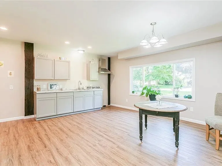 Bright open living space with a small kitchenette, a round dining table with a plant, large window overlooking greenery, and a wooden floor.