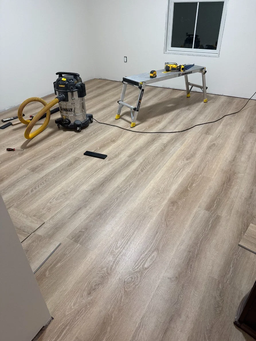 Room with light wood laminate flooring, construction tools including a wet/dry vacuum, a cordless drill, tape measure, a small plank, and a step ladder, during flooring installation.