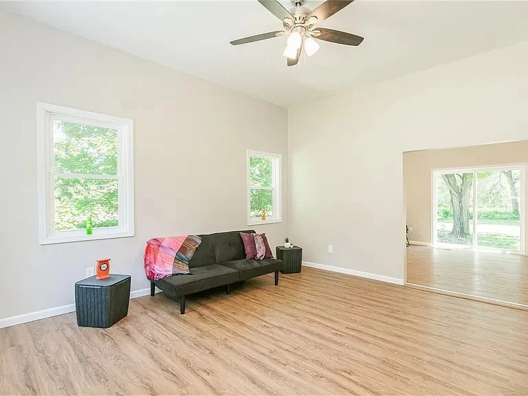 Living room with a black sofa, pink throw blanket, two windows showing green trees outside, hardwood floors, ceiling fan, and a doorway leading to another room with sliding glass doors.