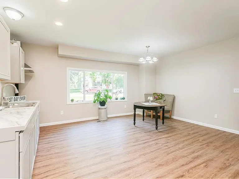Empty living room with large window, small table with chair, and kitchen area with white cabinets and marble countertop.