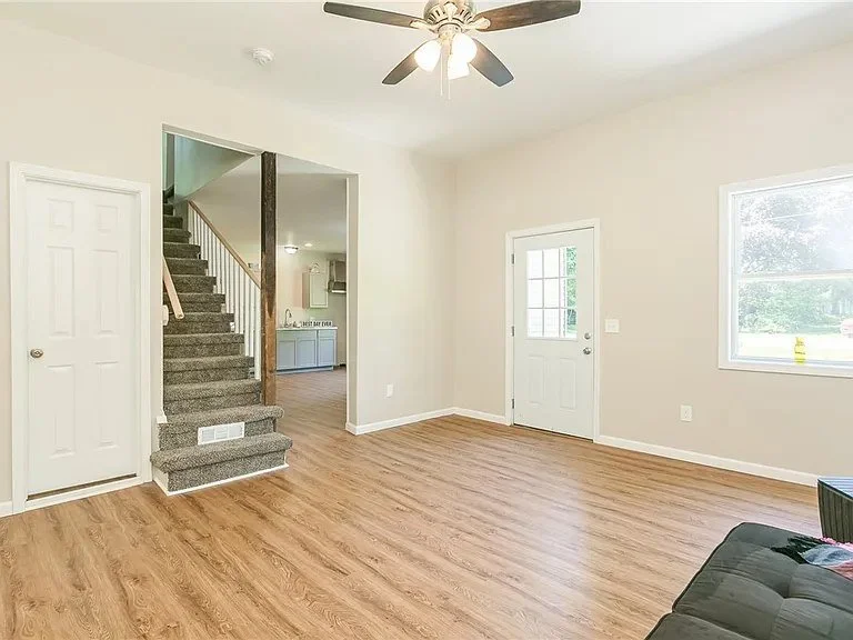 Empty living room with wooden floors, a ceiling fan, a window, a door leading outside, and a staircase leading upstairs.