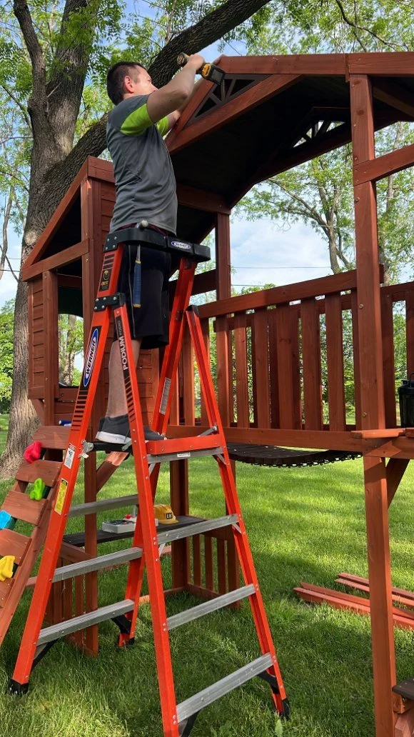 A man working on building a wooden playhouse or treehouse in a backyard, standing on an orange ladder and using a power drill, with trees and a grassy area in the background.